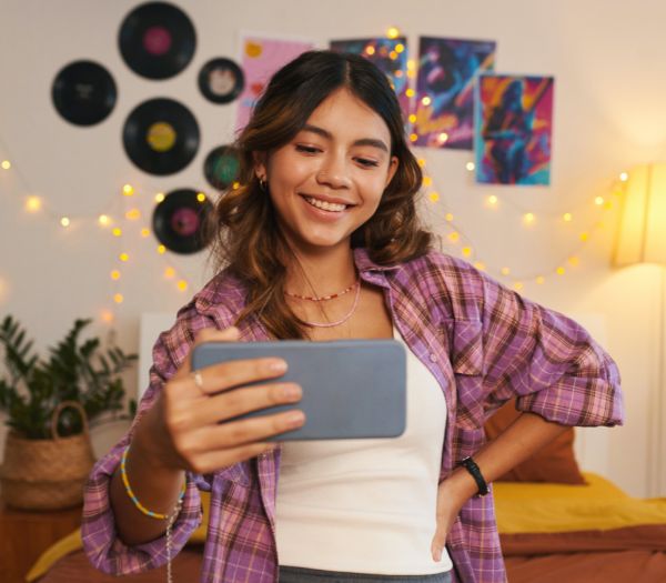 Student smiles while taking a selfie with a smartphone in a decorated dorm room.